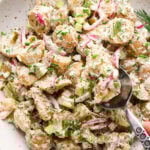 A large shallow speckled bowl filled with creamy mayo-based dill pickle potato salad with a spoon lifting out a portion to show the texture. The potato salad is garnished with fresh dill, fresh snipped chives, and black pepper.