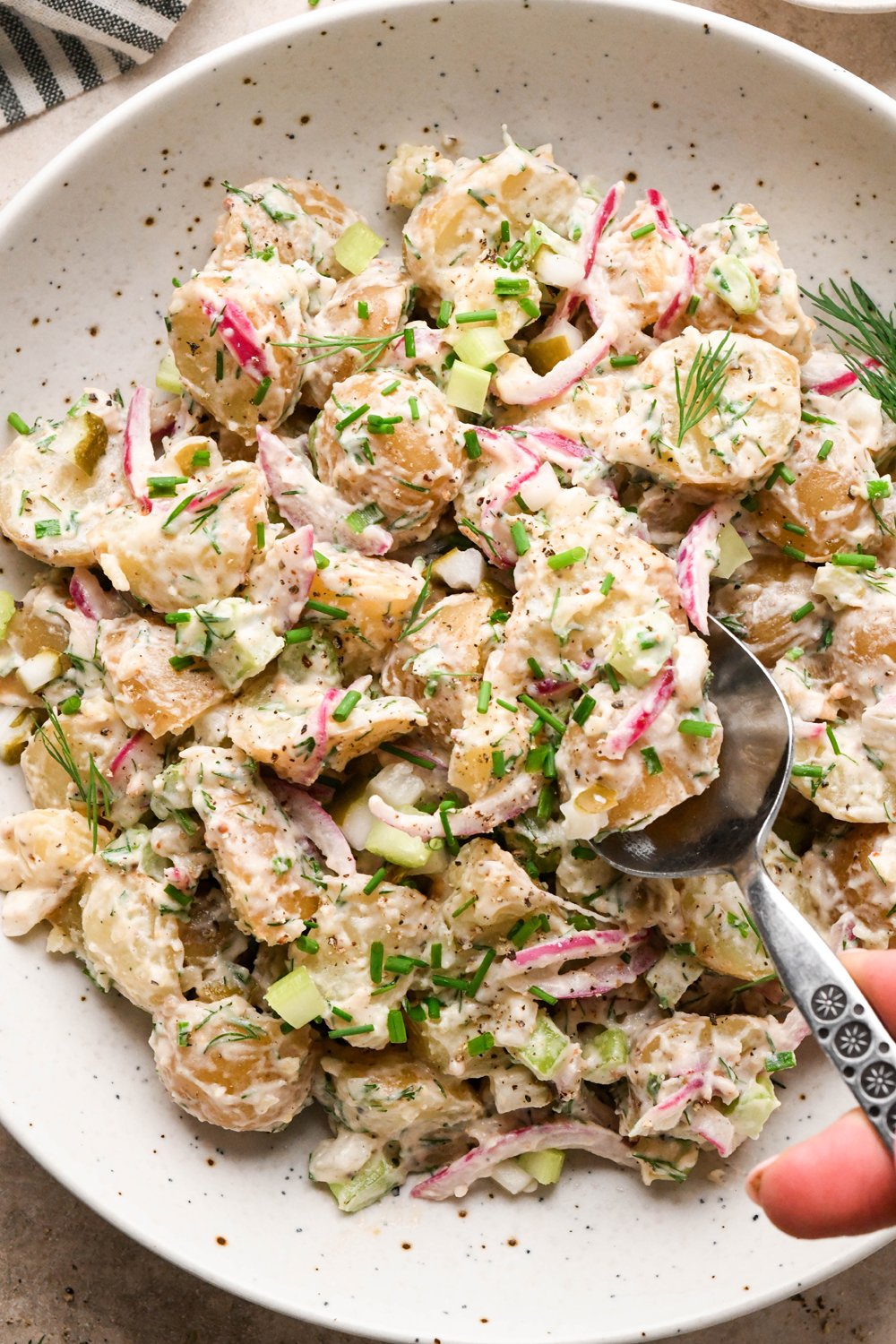A large shallow speckled bowl filled with creamy mayo-based dill pickle potato salad with a spoon lifting out a portion to show the texture. The potato salad is garnished with fresh dill, fresh snipped chives, and black pepper.
