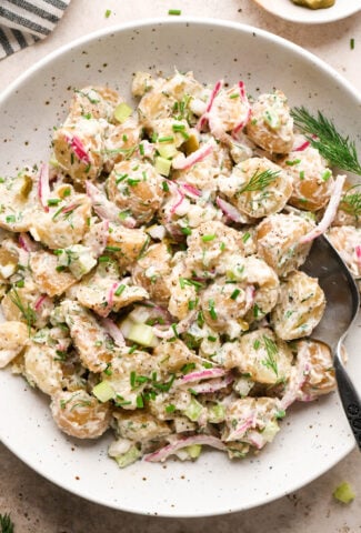 A large shallow speckled bowl filled with creamy mayo-based dill pickle potato salad. A spoon is angled into the bowl, and the potato salad is garnished with fresh dill, fresh snipped chives, and black pepper.