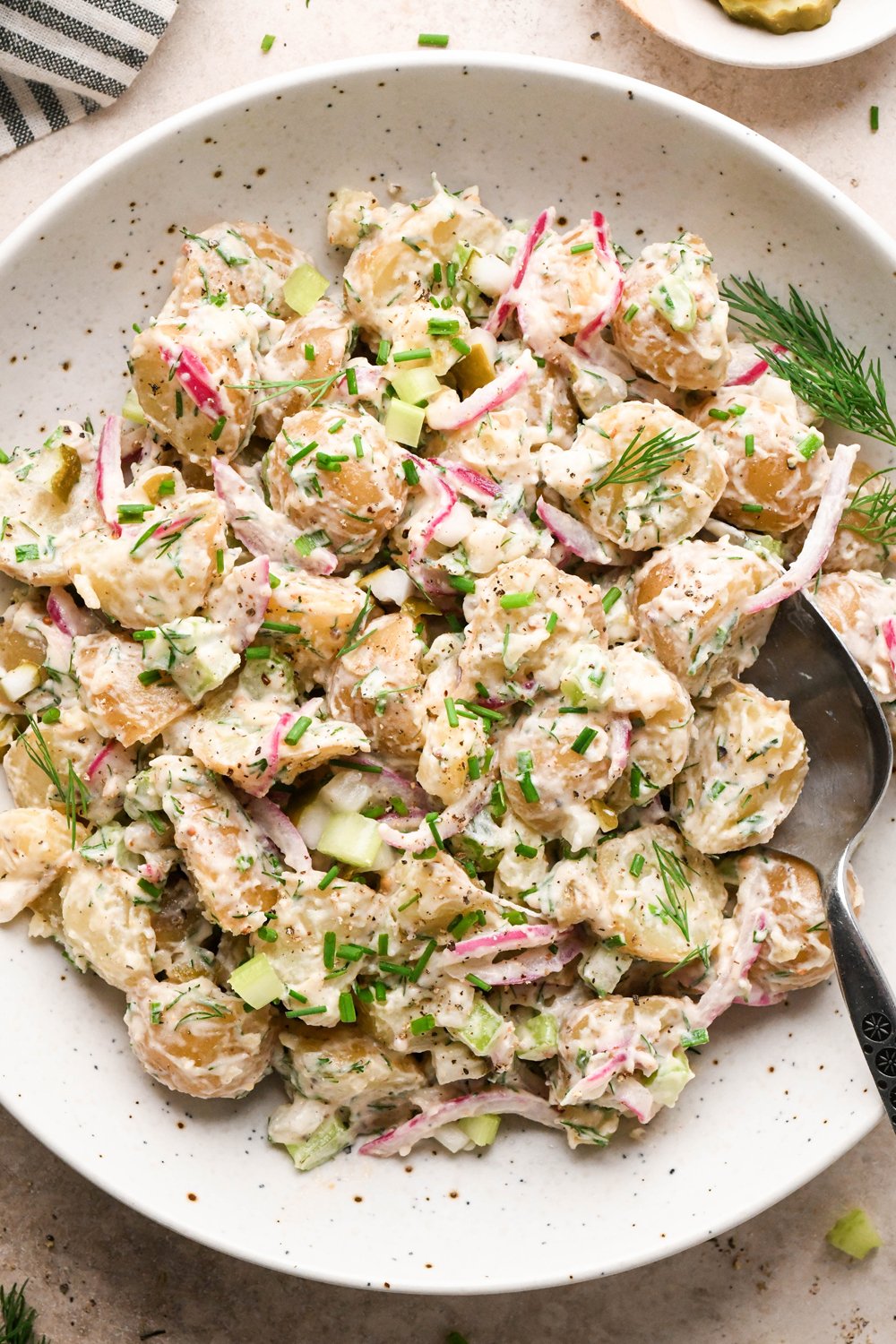 A large shallow speckled bowl filled with creamy mayo-based dill pickle potato salad. A spoon is angled into the bowl, and the potato salad is garnished with fresh dill, fresh snipped chives, and black pepper.