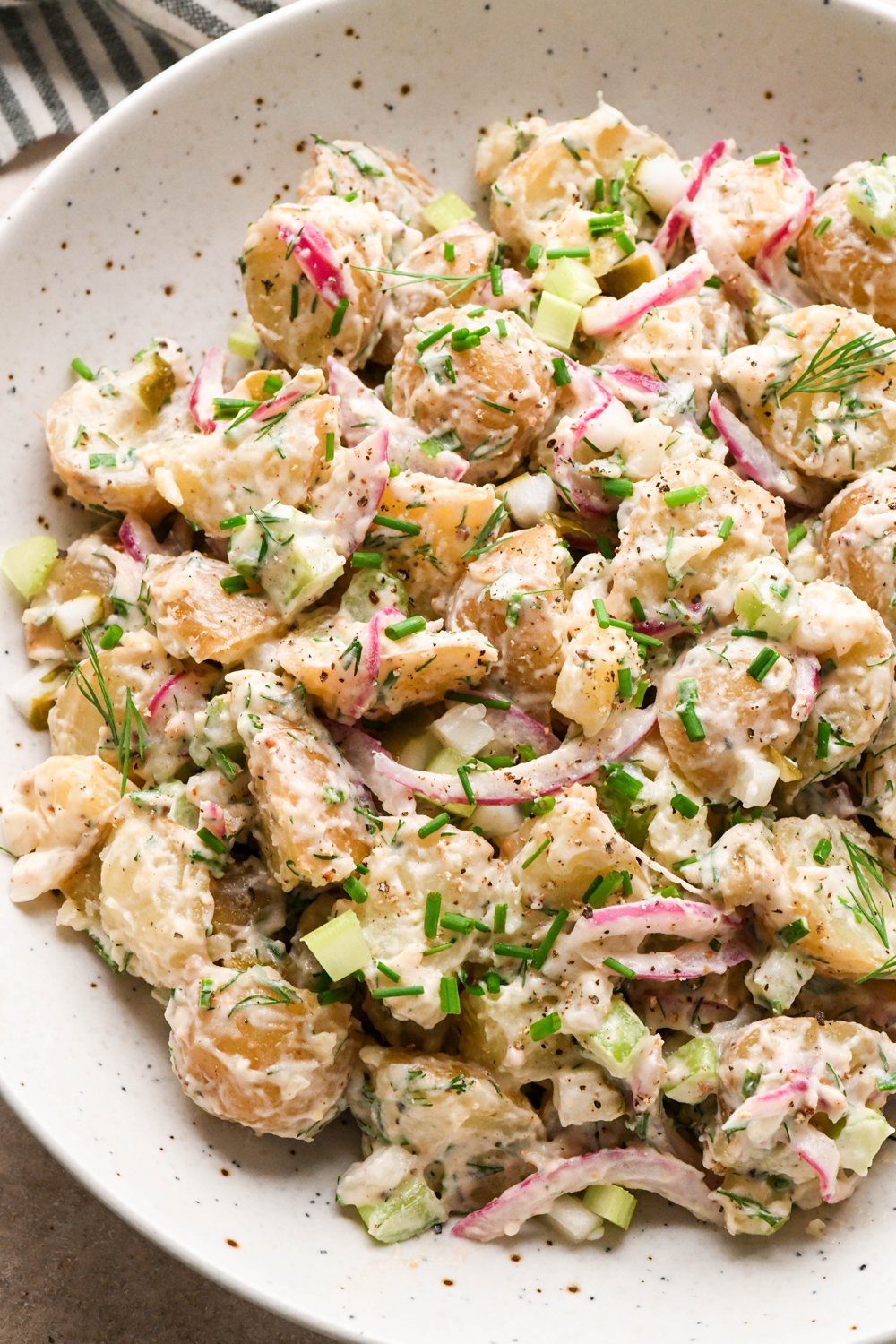 A large shallow speckled bowl filled with creamy mayo-based dill pickle potato salad. The potato salad is garnished with fresh dill, fresh snipped chives, and black pepper.
