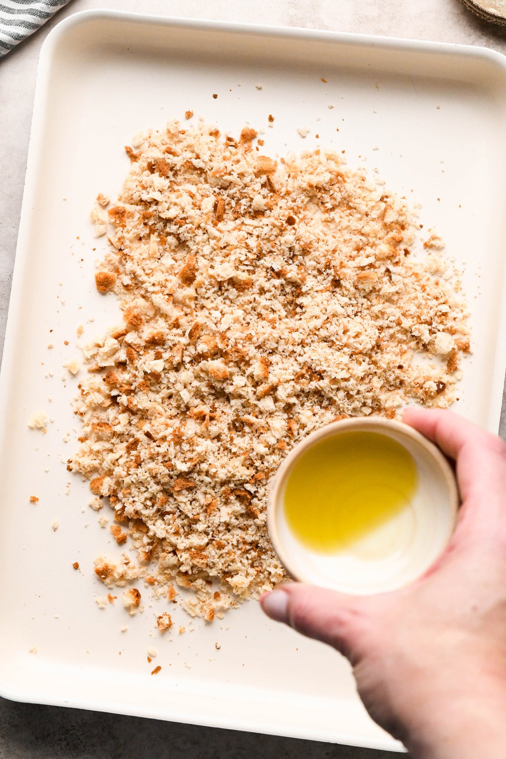 How to make a Caesar Salad with gluten free breadcrumbs: Breadcrumbs on a white baking sheet, a hand is pouring olive oil from a small dish onto the crumbs.