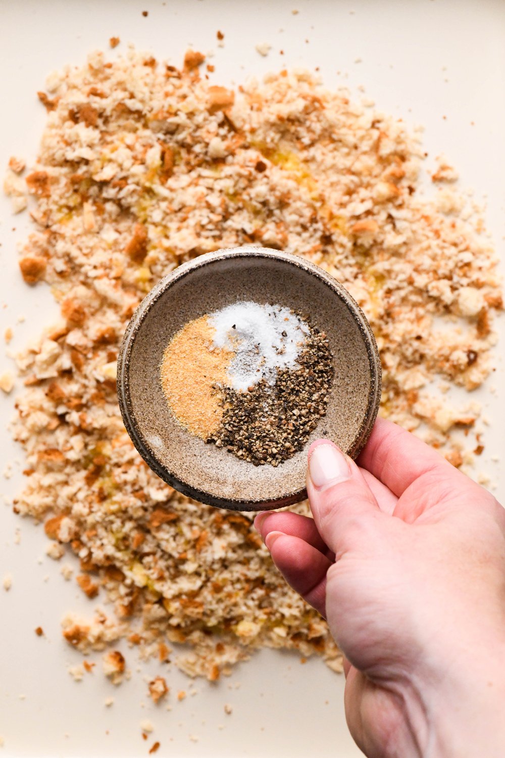 How to make a Caesar Salad with gluten free breadcrumbs: A hand holding a small dish with salt, pepper, and garlic powder over the breadcrumbs on a white baking sheet. 