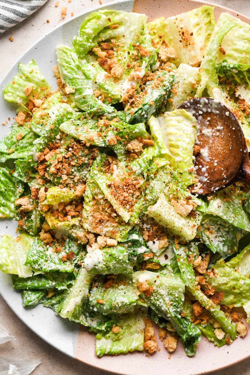 A large ceramic serving platter with a Caesar salad tossed in dressing and topped with grated parmesan, and toasty gluten free breadcrumbs. A wooden serving spoon is angled into the salad to lift out a portion.
