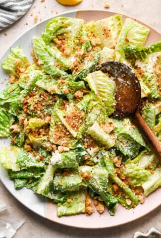 A large ceramic serving platter with a Caesar salad tossed in dressing and topped with grated parmesan, and toasty gluten free breadcrumbs. A wooden serving spoon is angled into the salad to lift out a portion.