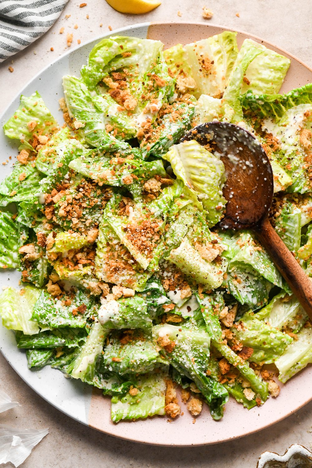 A large ceramic serving platter with a Caesar salad tossed in dressing and topped with grated parmesan, and toasty gluten free breadcrumbs. A wooden serving spoon is angled into the salad to lift out a portion.