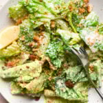 A small serving bowl of Caesar salad with gluten free breadcrumbs and grated parmsan. There is a wedge of lemon in the bowl and a fork is angled in with a bite of salad on it that's coated in creamy dressing.