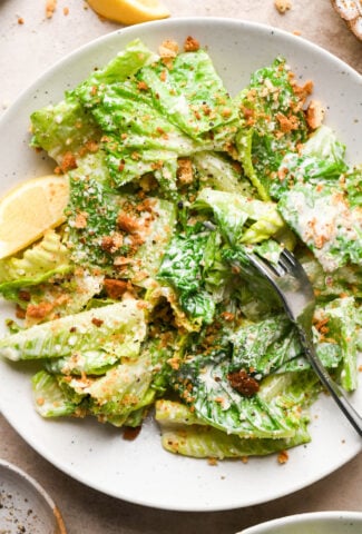 A small serving bowl of Caesar salad with gluten free breadcrumbs and grated parmsan. There is a wedge of lemon in the bowl and a fork is angled in with a bite of salad on it that's coated in creamy dressing.