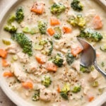 A shallow cream colored ceramic bowl of creamy dairy free chicken and broccoli soup on a light brown background. A spoon is digging into the middle of the soup for a bite, and a striped napkin is next to the bowl.