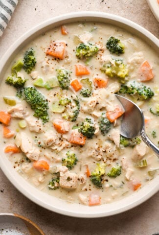 A shallow cream colored ceramic bowl of creamy dairy free chicken and broccoli soup on a light brown background. A spoon is digging into the middle of the soup for a bite, and a striped napkin is next to the bowl.