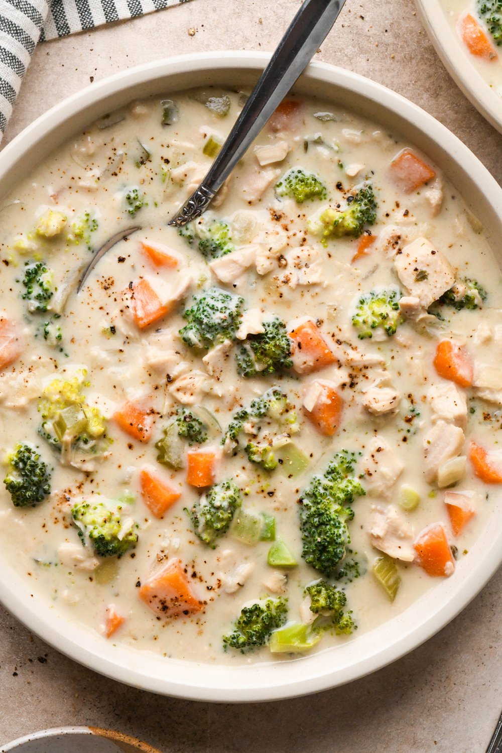 A shallow cream colored ceramic bowl of creamy dairy free chicken and broccoli soup on a light brown background. A spoon is tucked into the edge of the bowl and a striped napkin is next to it.