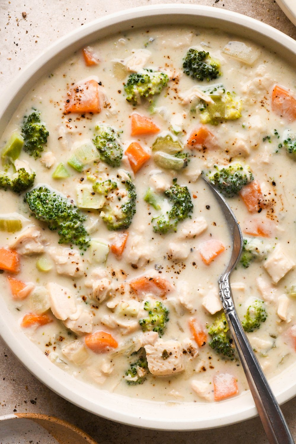 A shallow cream colored ceramic bowl of creamy dairy free chicken and broccoli soup on a light brown background. A spoon is tucked into the edge of the bowl.