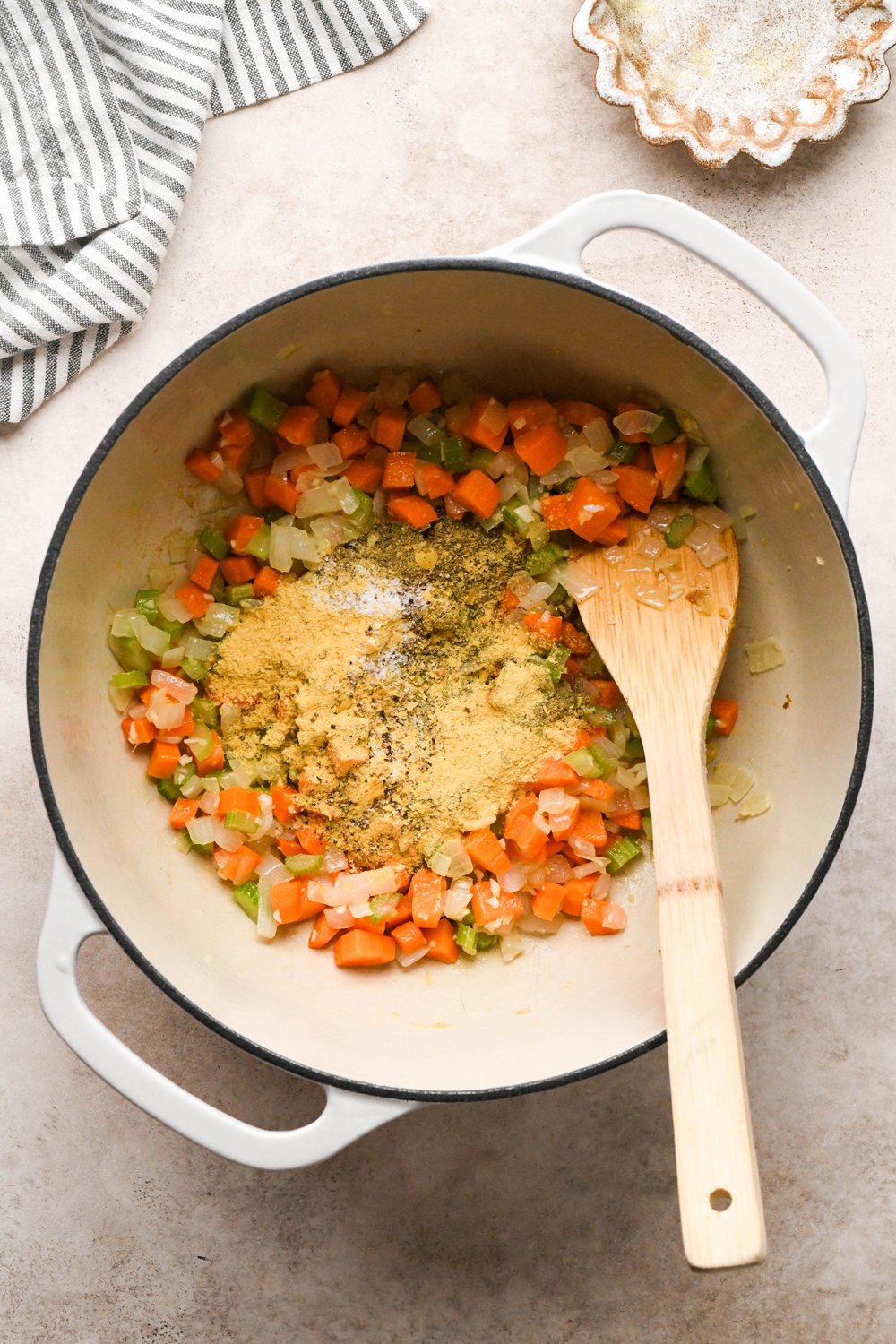 How to make Chicken and Broccoli Soup: Diced carrots, diced celery, diced onion, and chopped garlic in a large off-white ceramic soup pot with olive oil, after sautéing. All purpose seasoning, nutritional yeast, salt, and pepper are on top of the softened veggies. 