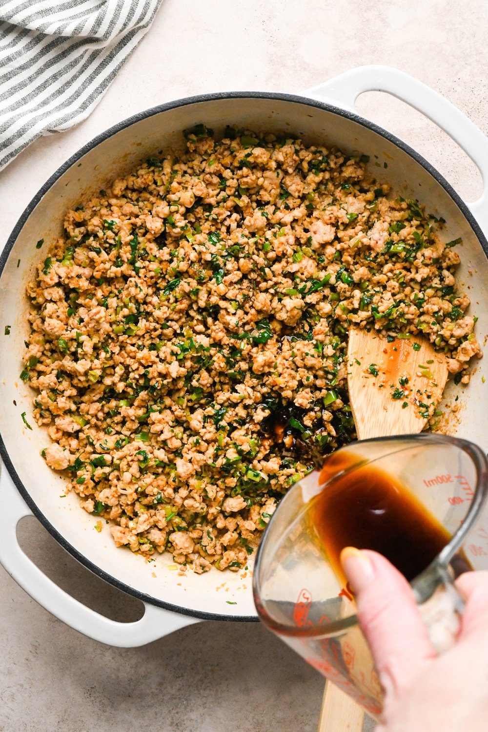 How to make garlic scallion noodles: A hand pouring the coconut amino, tamari, and vinegar mixture into the skillet with the cooked ground chicken.