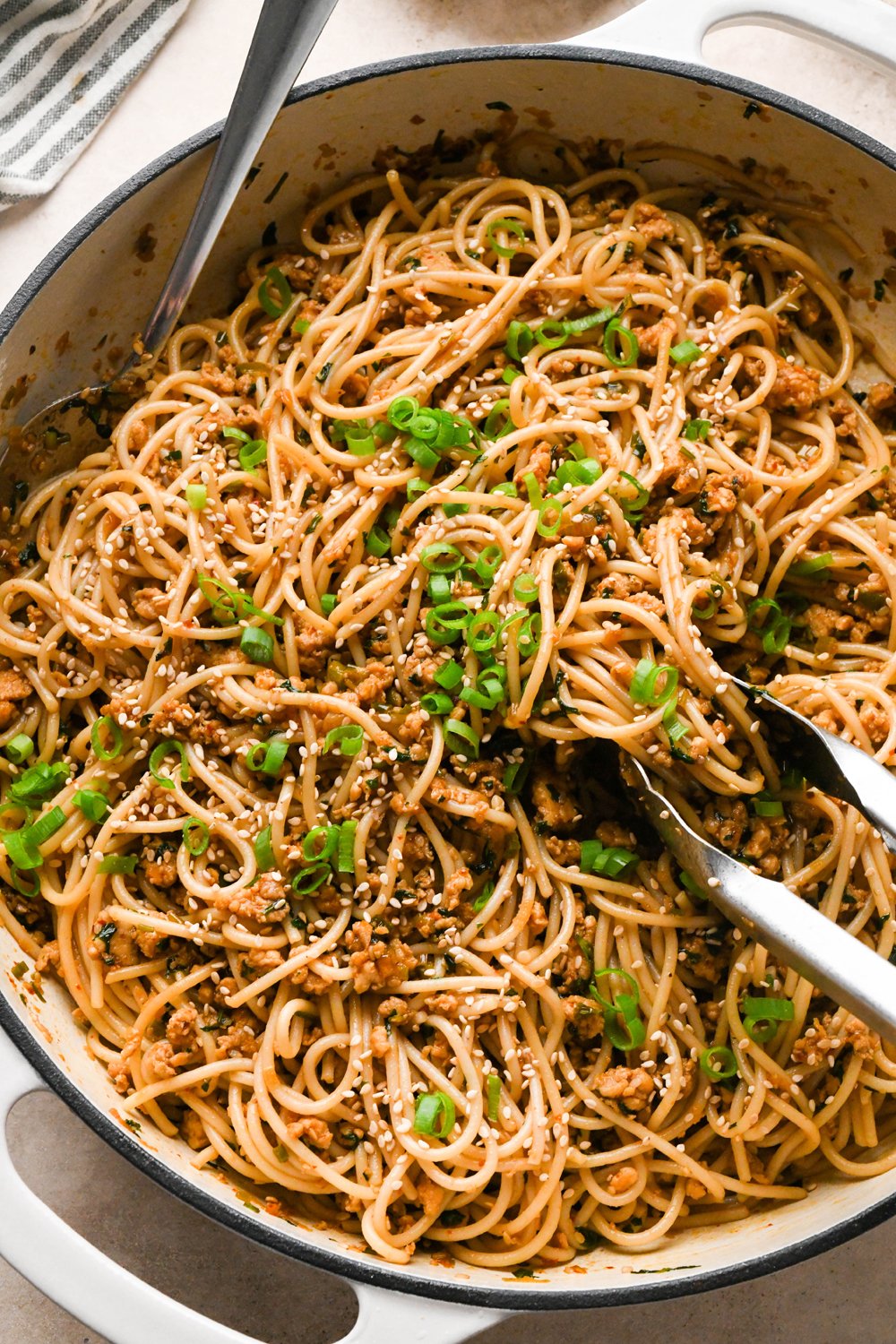 Prepared garlic scallion noodles in a large shallow ceramic skillet, garnished with sesame seeds and finely sliced scallions. A pair of metal tongs is angled into the noodles, and a large metal spoon is tucked into the side of the skillet.