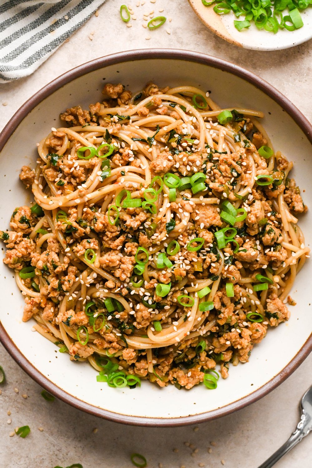 A large creamy colored speckled ceramic bowl filled with garlic scallion noodles, garnished with sesame seeds and thinly sliced scallions.
