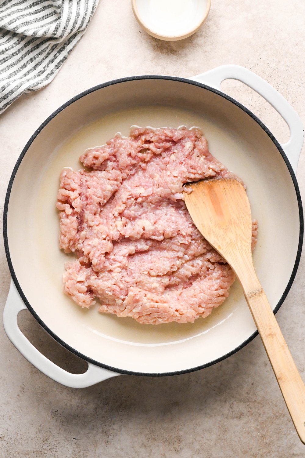 How to make garlic scallion noodles: Raw ground chicken in a large cream colored enamled skillet, chicken is pressed into a thin layer to cook.