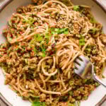 A large creamy colored speckled ceramic bowl filled with garlic scallion noodles, a fork angled into the noodles with a bite ready be lifted out of the bowl. The noodles are garnished with sesame seeds and thinly sliced scallions.