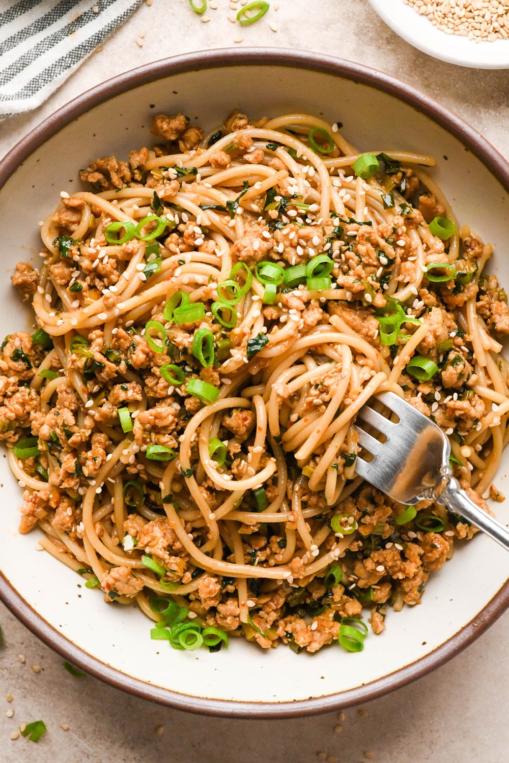 A large creamy colored speckled ceramic bowl filled with garlic scallion noodles, a fork angled into the noodles with a bite ready be lifted out of the bowl. The noodles are garnished with sesame seeds and thinly sliced scallions.
