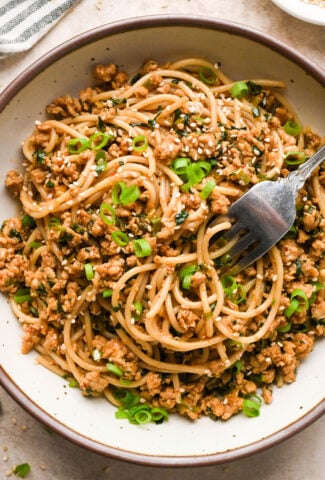 A large creamy colored speckled ceramic bowl filled with garlic scallion noodles, a fork is angled into the noodles. The noodles are garnished with sesame seeds and thinly sliced scallions.