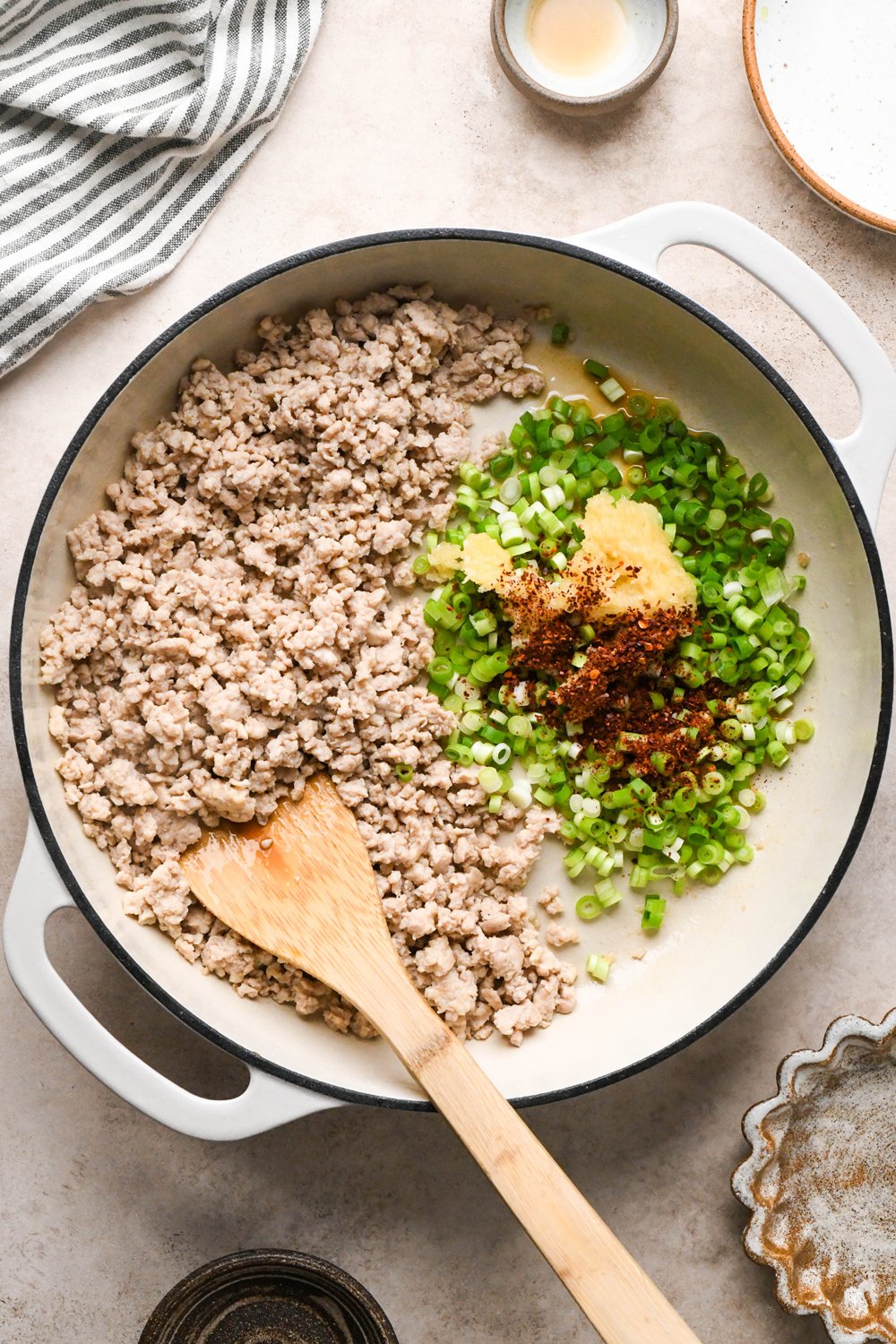 How to make garlic scallion noodles: Ground chicken broken apart and cooked in a cream colored enameled skillet. Cooked chicken is pushed to one half of the skillet, and green onions, garlic, and chili flakes are added to the other side of the pan.