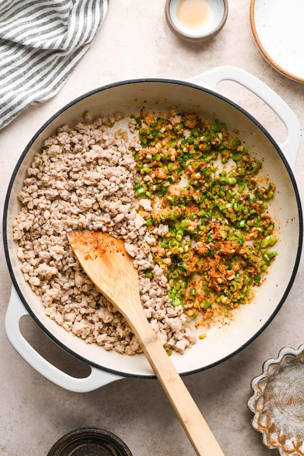 How to make garlic scallion noodles: Ground chicken broken apart and cooked in a cream colored enameled skillet. Cooked chicken is pushed to one half of the skillet, and green onions, garlic, and chili flakes have been briefly sautéed to soften the scallions on the other side of the pan.