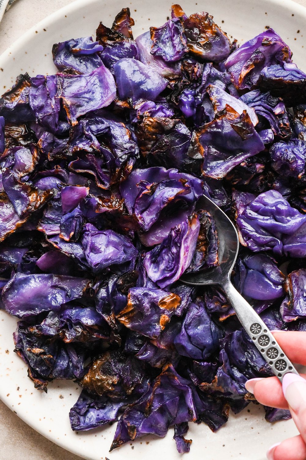 Pieces of roasted red cabbage on a large speckled plate, with a metal serving spoon lifting out a portion of the caramelized roasted veggie.