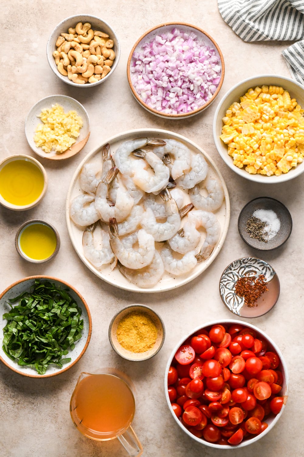 Overhead view of raw shrimp on a plate, surrounded by bowls of chopped cherry tomatoes, corn, raw cashews, olive oil, fresh basil, diced shallots, minced garlic, and a cup of broth on a light surface.