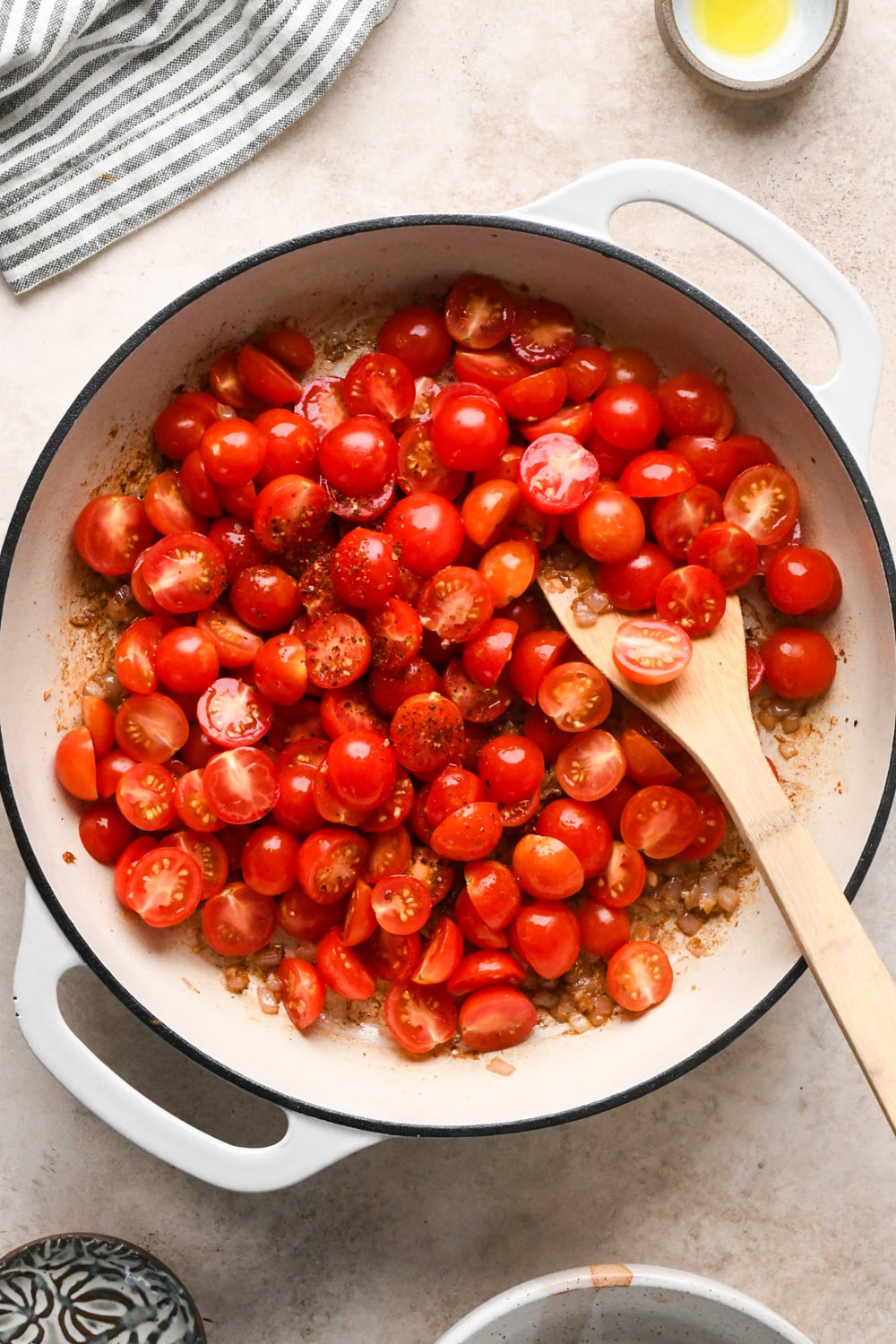 A white skillet filled with halved cherry tomatoes sprinkled with chili flakes. On top of the cooked shallots and garlic, and stirred with a wooden spoon. A striped towel and small bowls are nearby.
