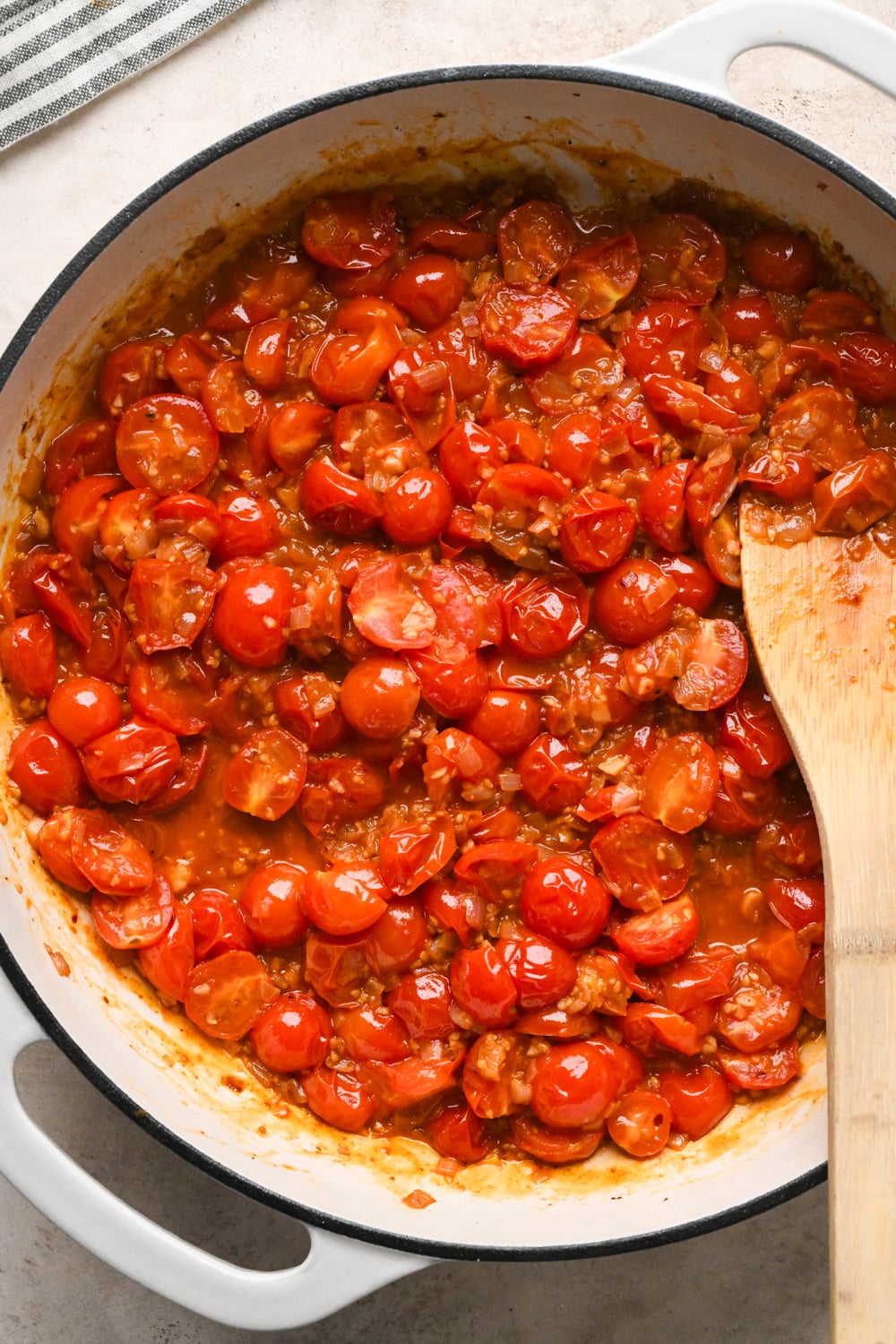 A white skillet filled with cooked cherry tomatoes that are developing into a sauce, being stirred with a wooden spoon.