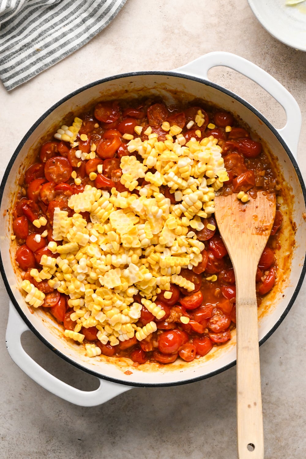 A white skillet filled with the cooked cherry tomatoes, fresh corn kernels added sits on a counter. A wooden spoon rests inside the pot, and a striped towel is nearby.