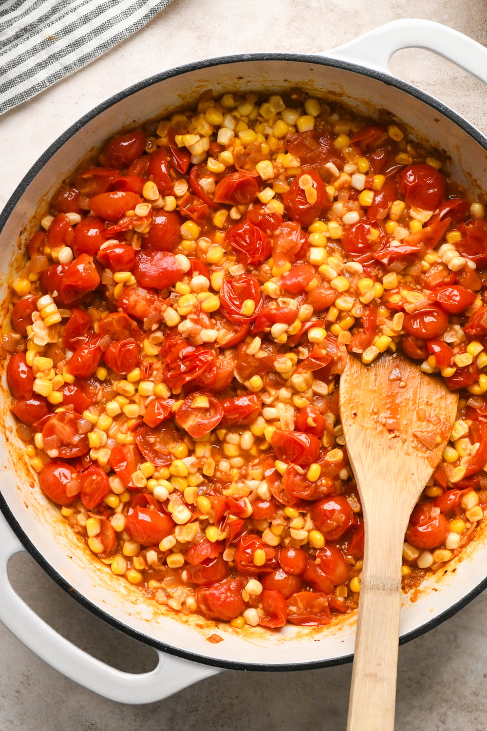 A white skillet filled with a cooked mixture of corn kernels and halved cherry tomatoes, stirred with a wooden spoon, sits on a light countertop.
