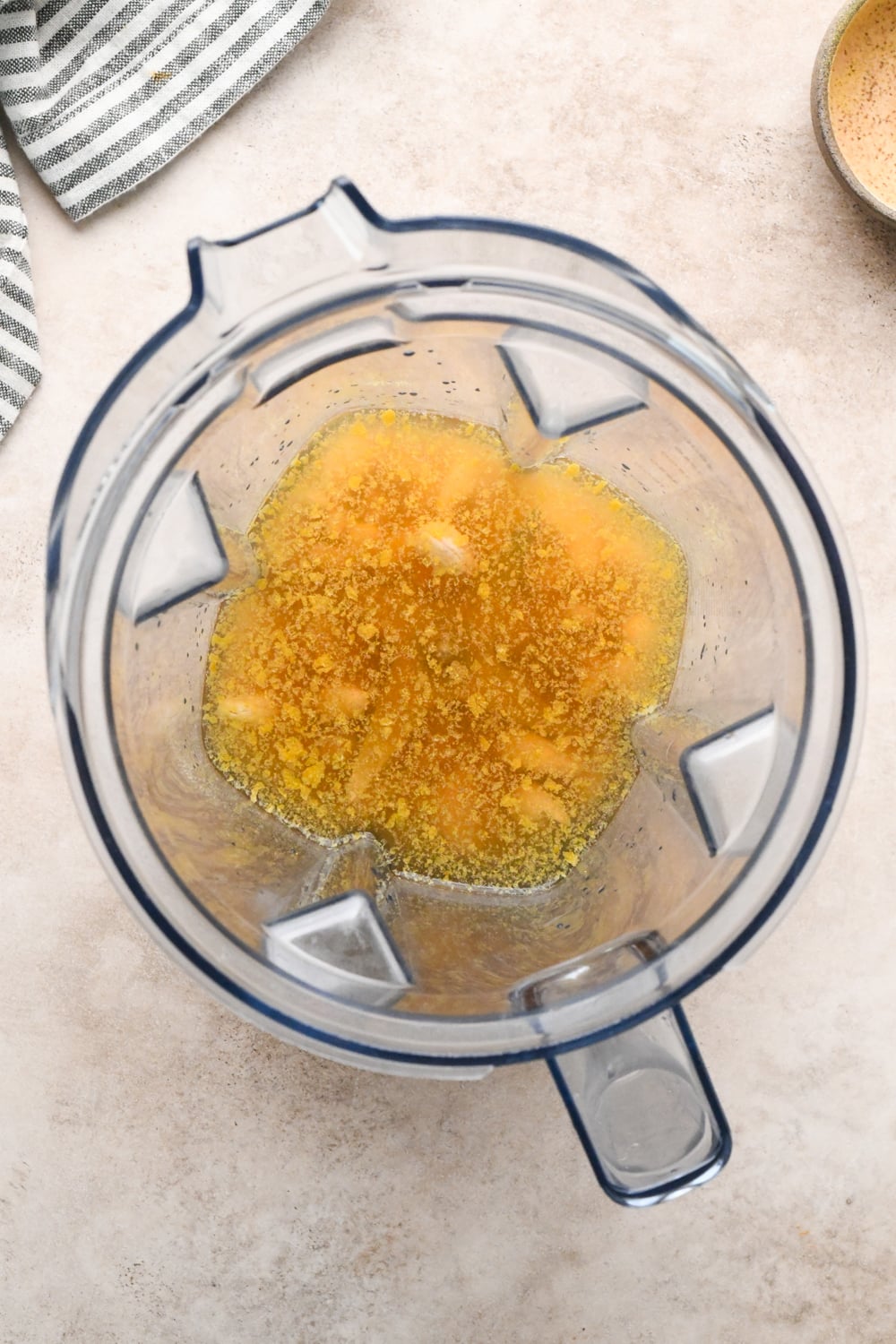Top-down view of a blender containing broth, nutritional yeast, and drained cashews before blending, sitting on a light-colored countertop with a striped cloth partially visible in the corner.