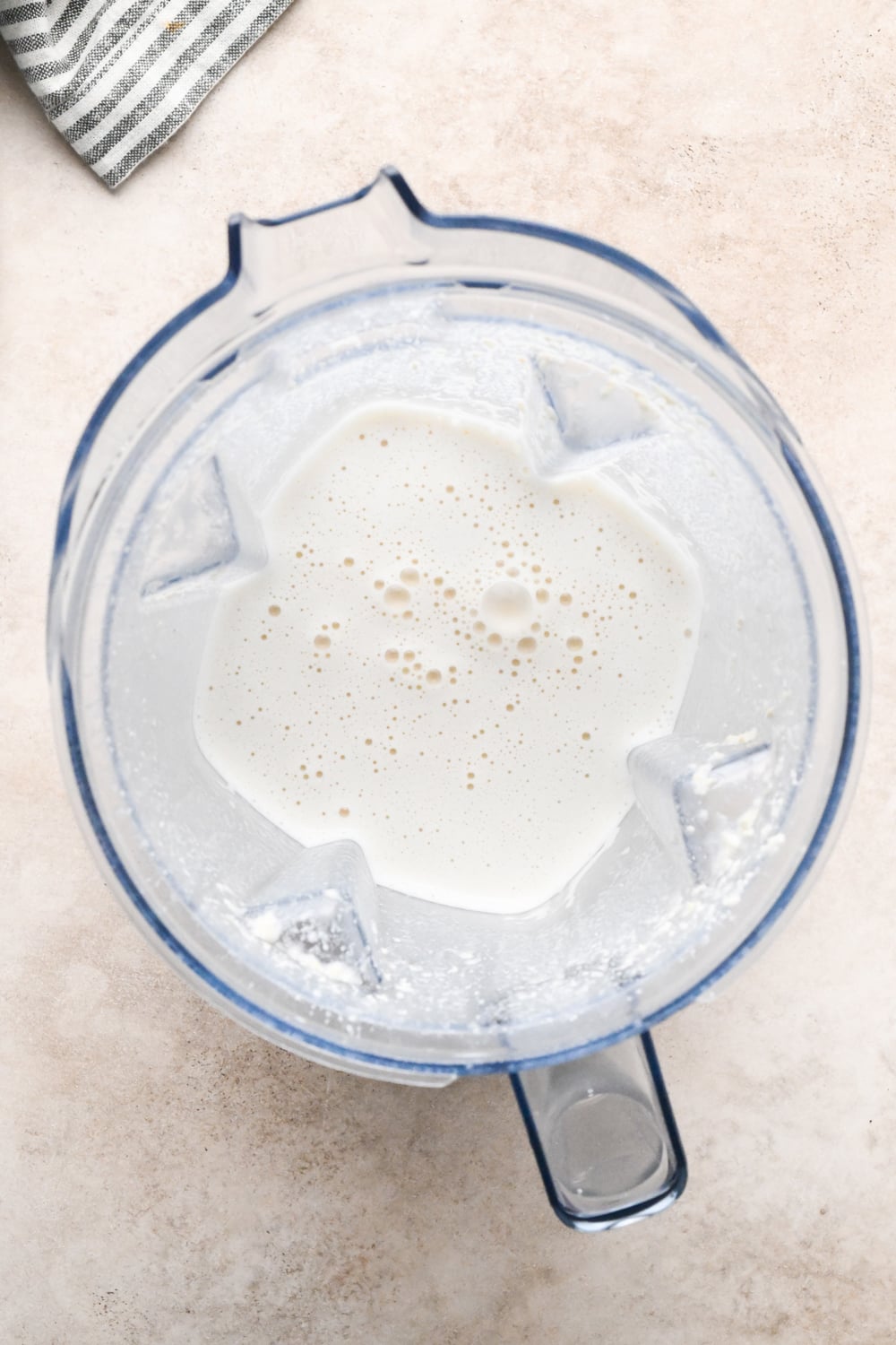 Top-down view of a blender containing the creamy blended mixture of broth, nutritional yeast, and drained cashews, sitting on a light-colored countertop with a striped cloth partially visible in the corner.