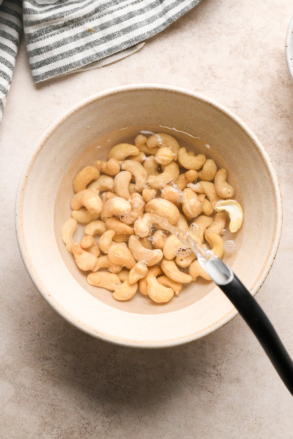 A bowl of raw cashews soaking in water, with a stream of water being poured in, on a light countertop next to a striped cloth.