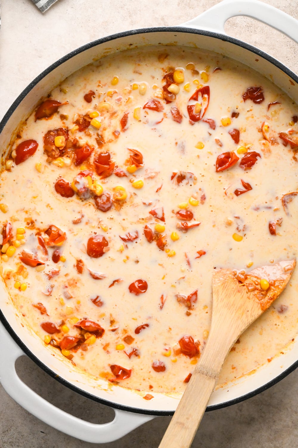 The white skillet just after adding the cashew cream mixture to the tomatoes and corn. A wooden spoon rests on the side, partially submerged in the thick, colorful mixture.
