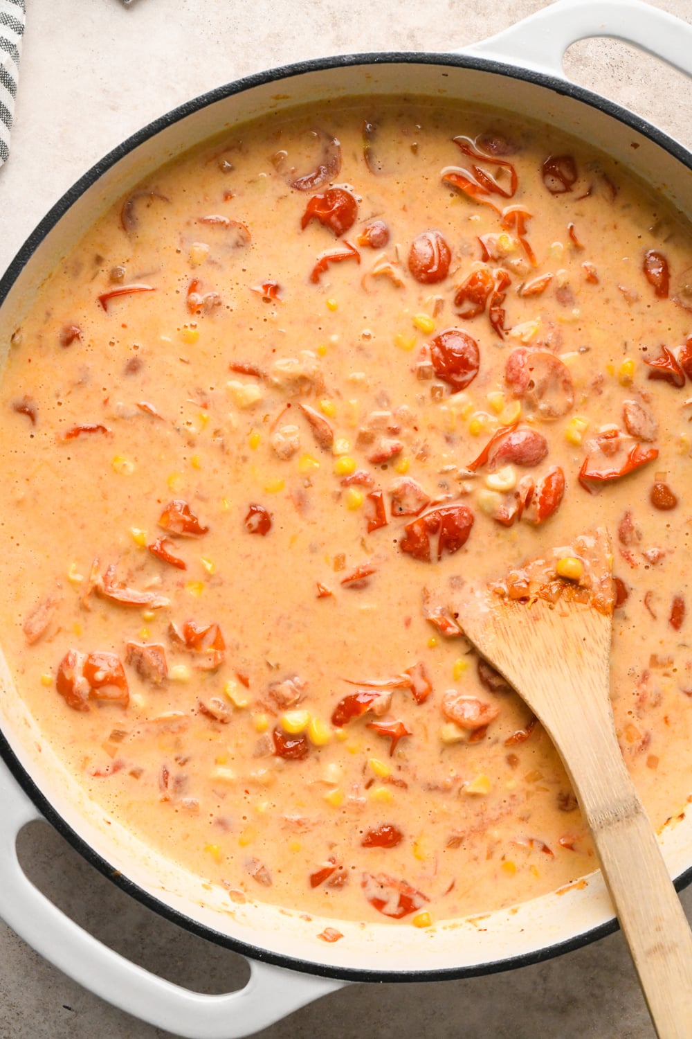 The white skillet after stirring the cashew cream mixture into the tomatoes and corn. A wooden spoon rests on the side, partially submerged in the thick, colorful mixture.