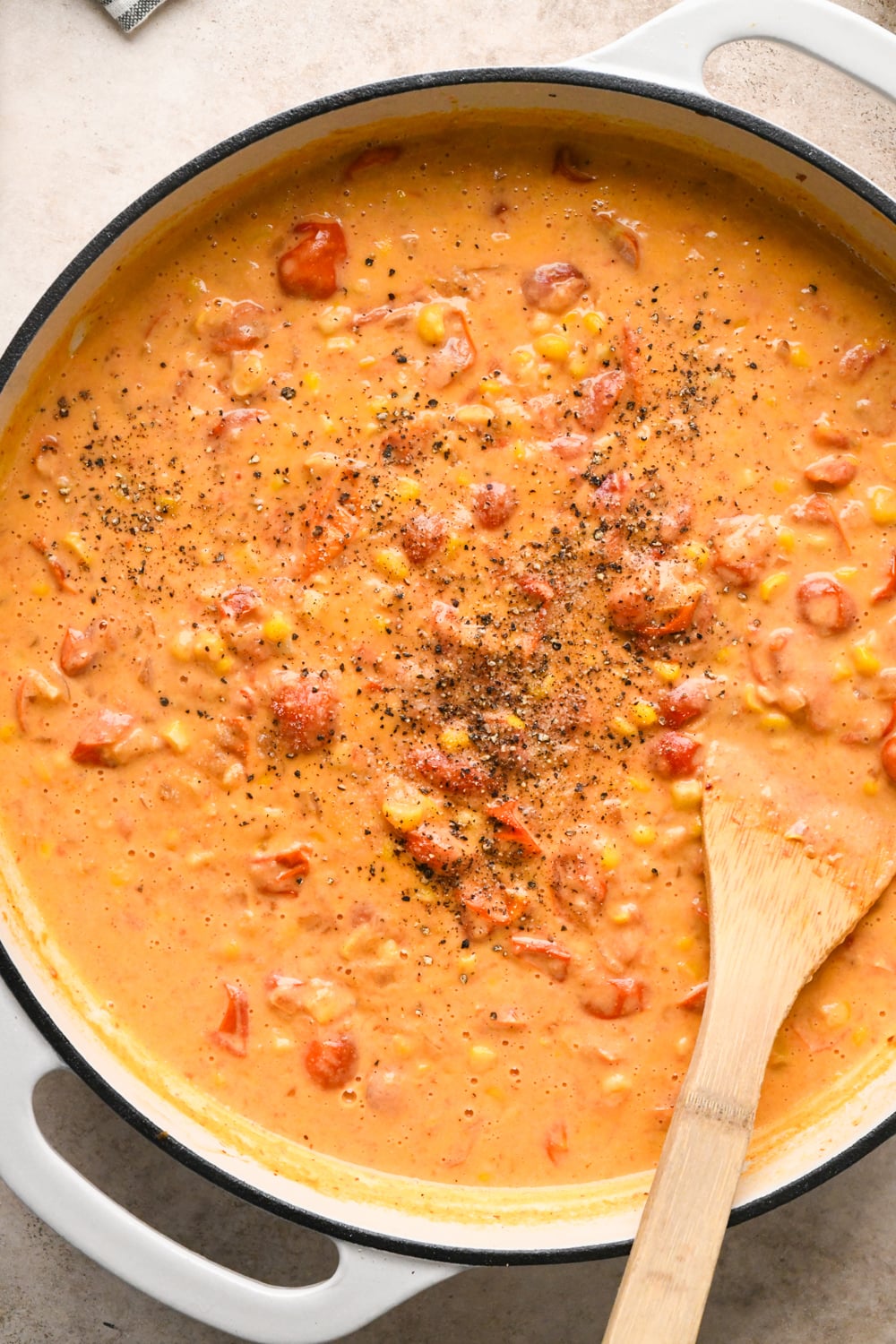 The white skillet after simmering the cashew cream into the sauce - the texture is thick and slightly darker red. Seasoned with black pepper and salt. A wooden spoon rests on the side, partially submerged in the thick, colorful mixture.