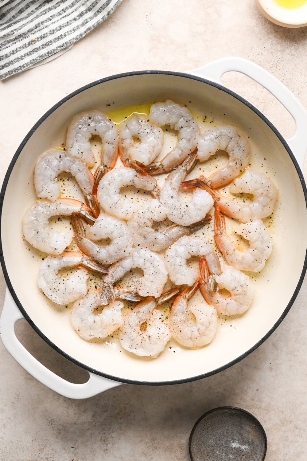Raw shrimp arranged in a circular pattern in a white skillet with olive oil, seasoned with salt and pepper, cooking on one side.