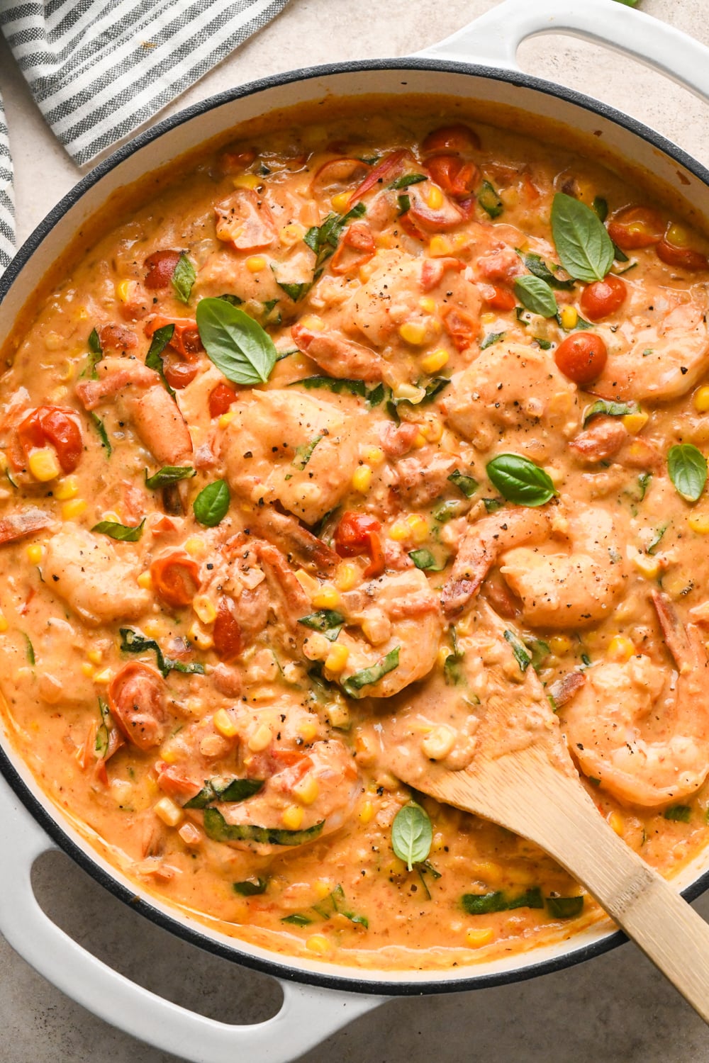 Creamy dairy-free shrimp and tomato skillet with sweet corn, cherry tomatoes, and basil, stirred with a wooden spoon in a white skillet.