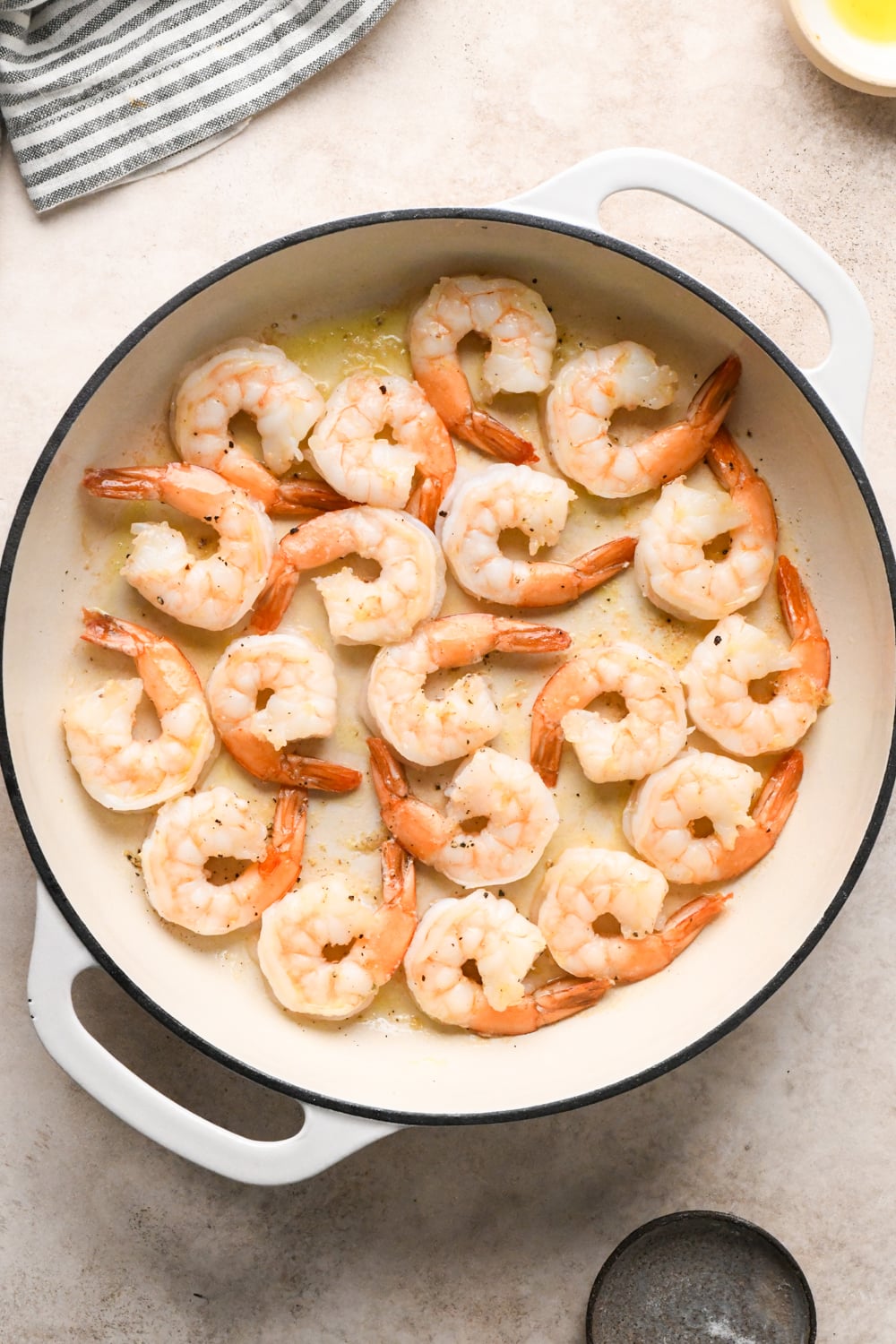 Cooked shrimp after flipping each one arranged in a white skillet with a light sauce, viewed from above. A striped towel and a small, empty bowl of olive oil are visible nearby on a light-colored surface.