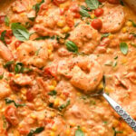 Creamy skillet dish with shrimp, cherry tomatoes, and corn, mixed in a rich, orange-colored cashew cream sauce. A spoon is partially dipped in, ready to serve. The skillet is garnished with tiny fresh basil leaves and a scatter of black pepper.