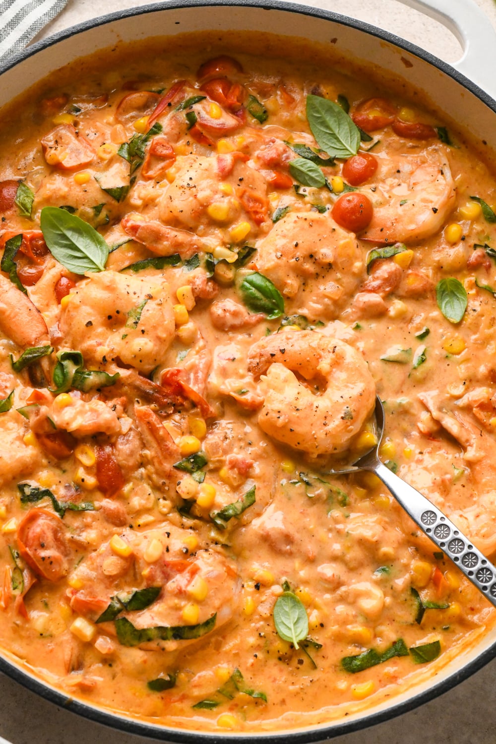 Creamy skillet dish with shrimp, cherry tomatoes, and corn, mixed in a rich, orange-colored cashew cream sauce. A spoon is partially dipped in, ready to serve. The skillet is garnished with tiny fresh basil leaves and a scatter of black pepper.