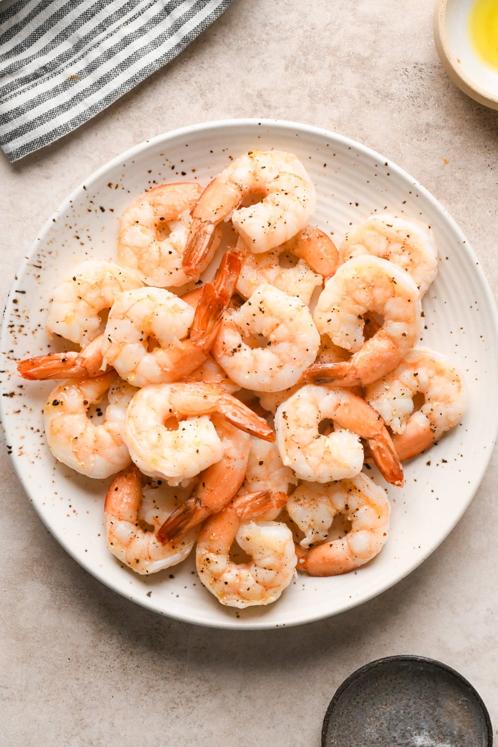 A white plate filled with cooked shrimp, lightly seasoned with pepper, sits on a light surface. Part of a striped towel and small dishes are visible nearby.