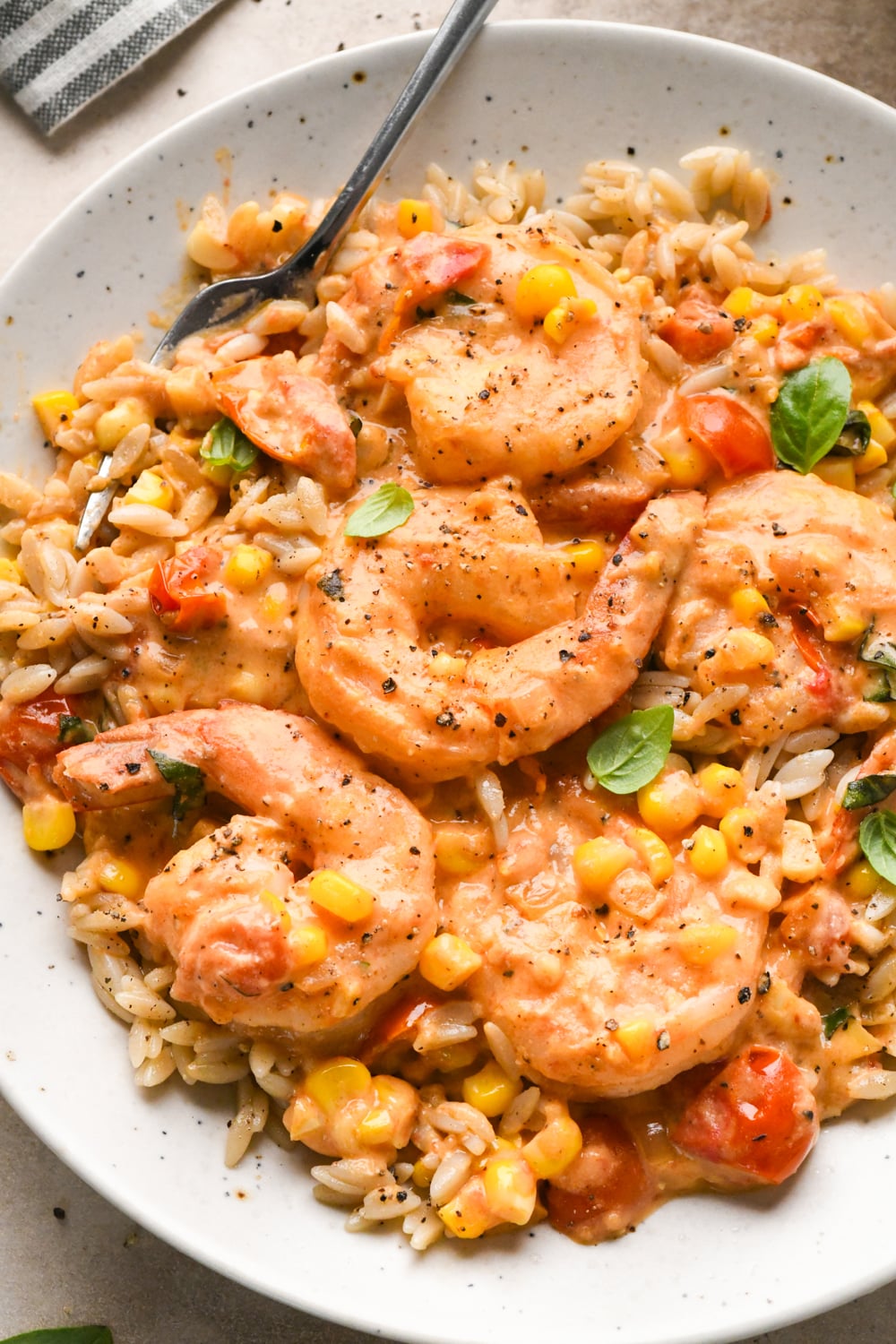A plate of creamy shrimp with corn, tomatoes, and herbs served over orzo pasta, garnished with fresh basil and black pepper. A fork rests on the edge of the dish.