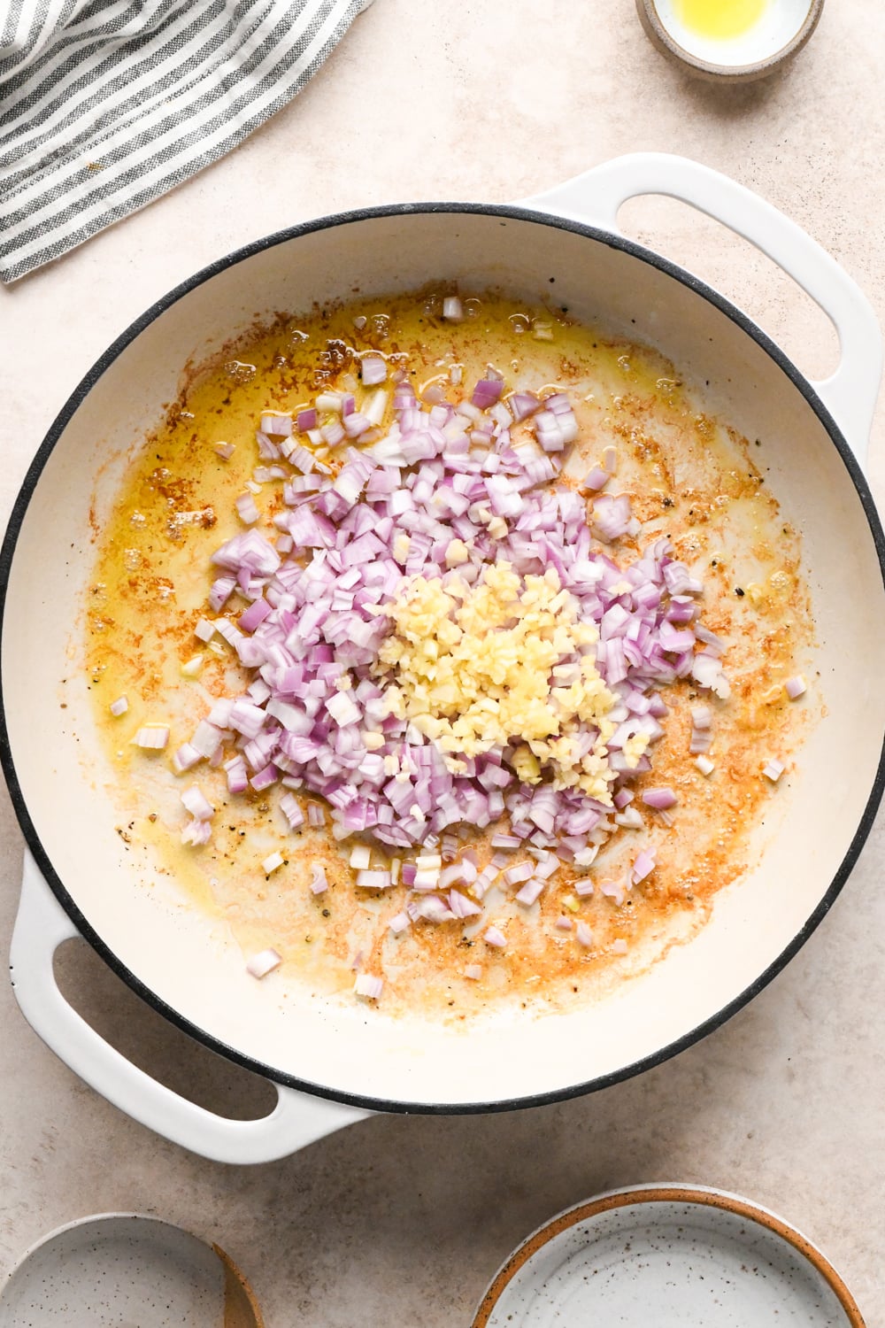 Chopped shallots and minced garlic in a white skillet with a light brown, caramelized base from cooking the shrimp. A small bowl of oil, a striped towel, and empty bowls are nearby on a light beige surface.