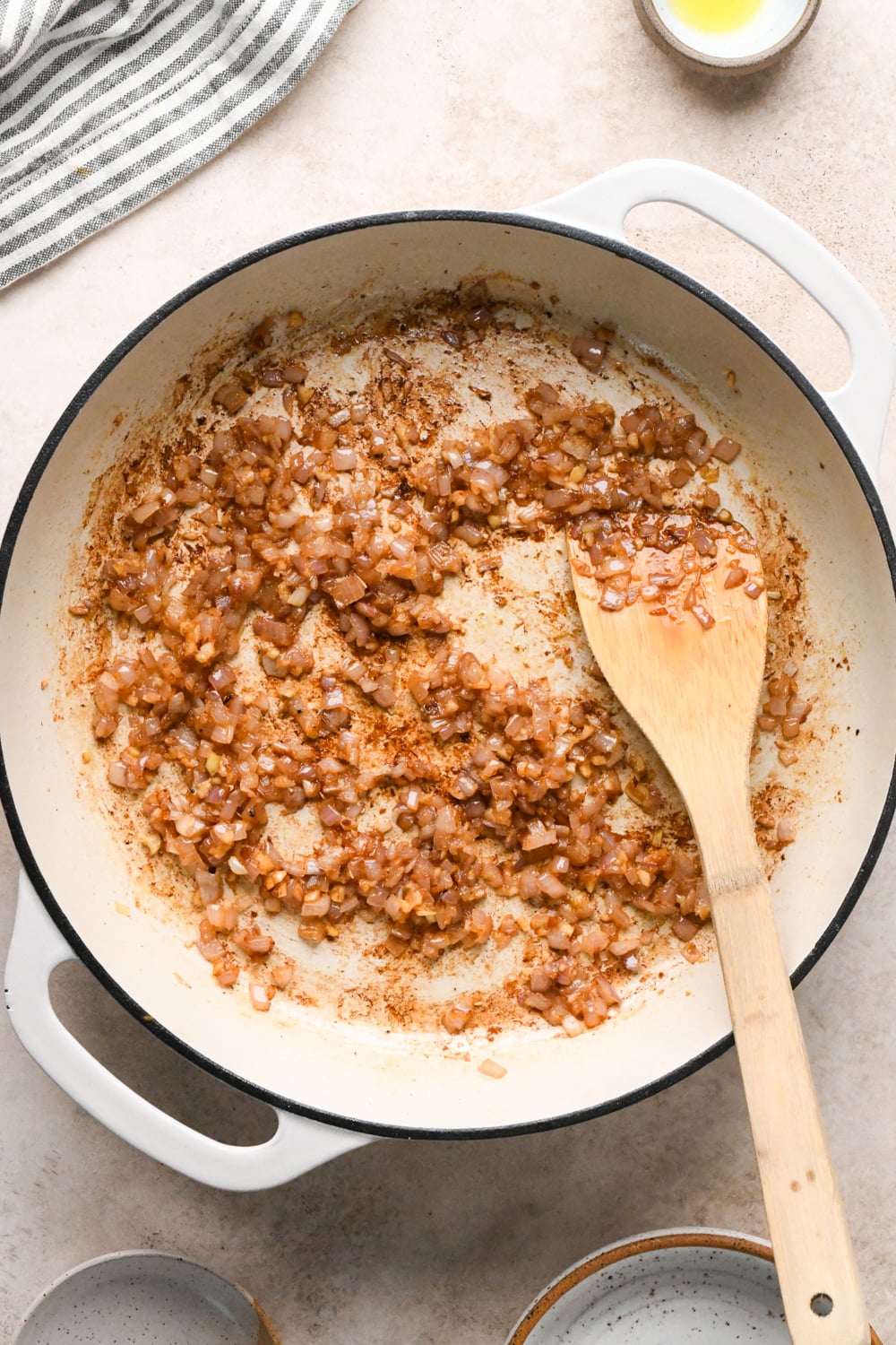 A white skillet with sautéed shallots and garlic, and browned bits on the bottom, stirred with a wooden spoon sits on a light countertop near a small bowl of oil and a striped towel.