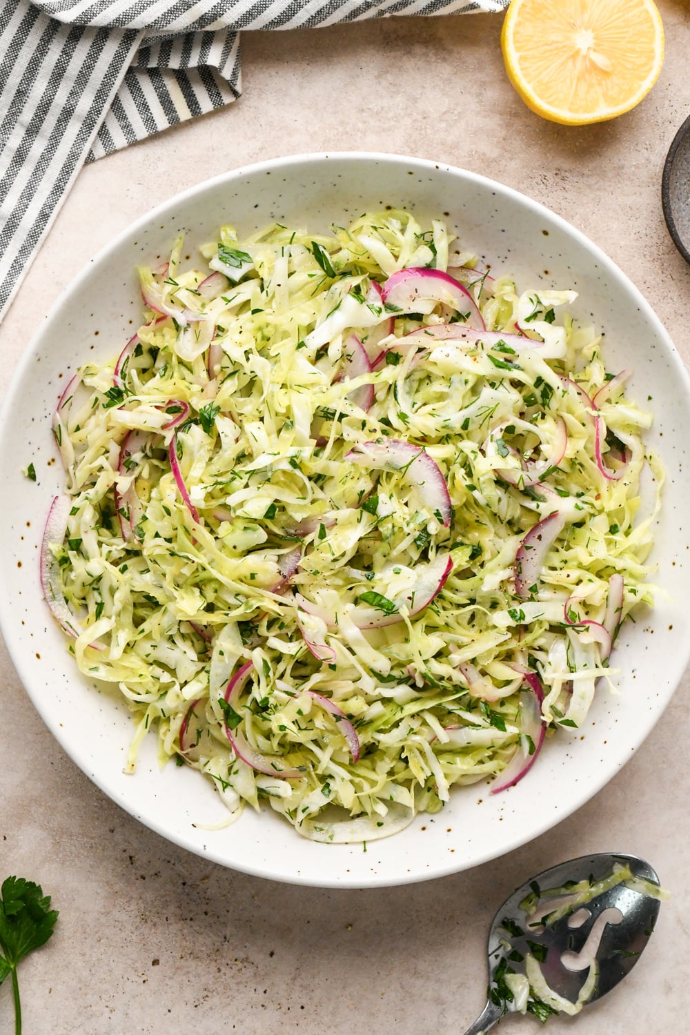 A white bowl filled with a shredded cabbage and fennel slaw with red onion, and fresh herbs, with a fork resting inside. A striped napkin and a lemon wedge are placed nearby on a light-colored surface.