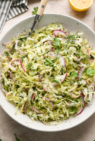 A white bowl filled with a shredded cabbage and fennel slaw with red onion, and fresh herbs, with a fork resting inside. A striped napkin and a lemon wedge are placed nearby on a light-colored surface.