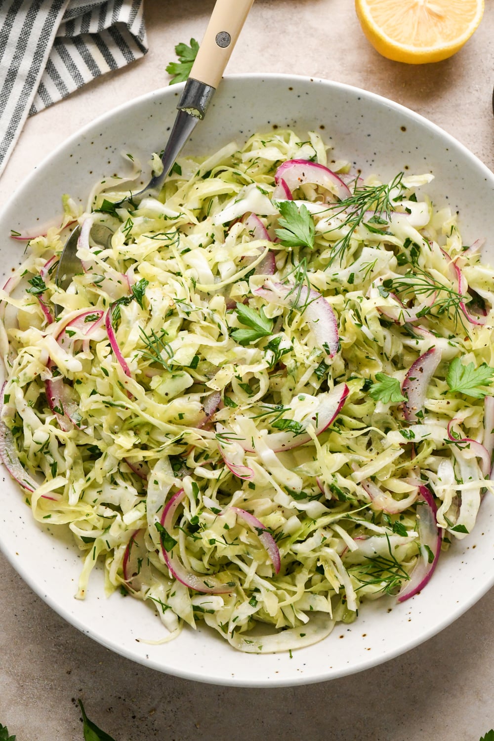 A white bowl filled with a shredded cabbage and fennel slaw with red onion, and fresh herbs, with a fork resting inside. A striped napkin and a lemon wedge are placed nearby on a light-colored surface.